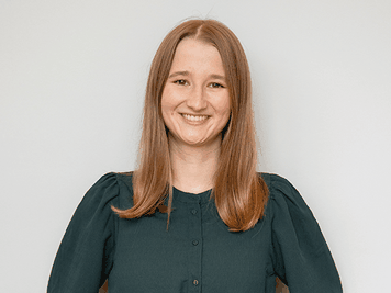 Uśmiechnięta dziewczyna z długimi brązowymi włosami, ubrana w ciemnozieloną bluzkę, stojąca na jasnym tle.