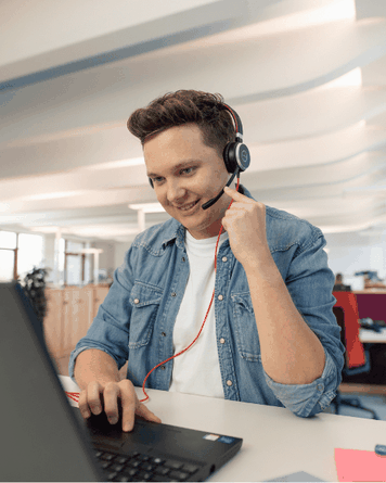 Młody mężczyzna w dżinsowej koszuli, noszący zestaw słuchawkowy, uśmiechnięty podczas pracy na laptopie w nowoczesnym biurze.