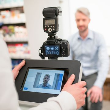 dm-Mitarbeiter, der einen sitzenden Mann mit einer Kamera und Blitzlicht fotografiert, während er Einstellungen an einem tabletähnlichen Gerät vornimmt. Regale im Hintergrund.