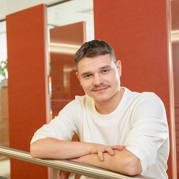 Eine Person mit kurzen Haaren und einem Schnurrbart, die ein weißes Hemd trägt, lehnt an einem Geländer in einem Raum mit roten Trennwänden und Pflanzen im Hintergrund.