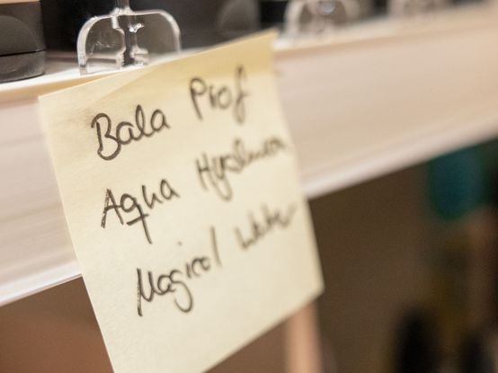 Nahaufnahme eines handgeschriebenen Zettels in einem Regal mit der Auflistung „Bala Prof, Aqua Hyaluron, Magical Water“ mit unscharfen Produkten im Hintergrund.