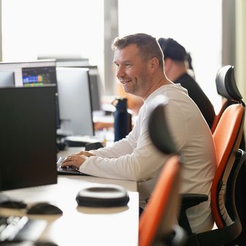 Mann im weißen Hemd lächelt, während er in einem Büro mit orangefarbenen Stühlen an einem Computer arbeitet. Eine weitere Person sitzt in der Nähe.