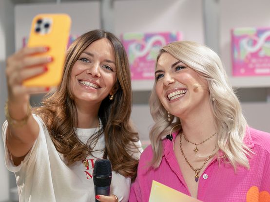 Zwei Frauen lächeln für ein Selfie; die eine hält ein Mikrofon, die andere bunte Moderationskarten. Sie befinden sich in einer hellen Umgebung mit rosa Dekoration.