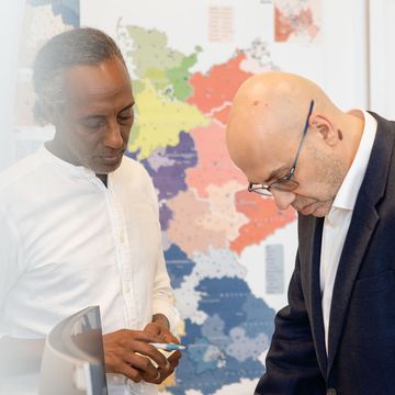 Zwei Männer diskutieren vor einer bunten Landkarte. Einer hält einen Stift in der Hand, und beide sind auf ein Dokument auf dem Tisch konzentriert.