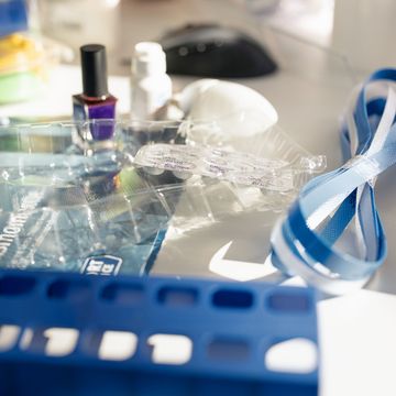 Ein überfüllter Schreibtisch mit einem Nagellackfläschchen, Augentropfenbehältern, einer Maus, einem Schlüsselband und einem blauen Plastikorganizer im Sonnenlicht.