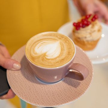Eine Person hält einen Milchkaffee in einer lilafarbenen Tasse mit Untertasse, während im Hintergrund ein mit roten Beeren gekröntes Törtchen auf einem weißen Teller liegt.