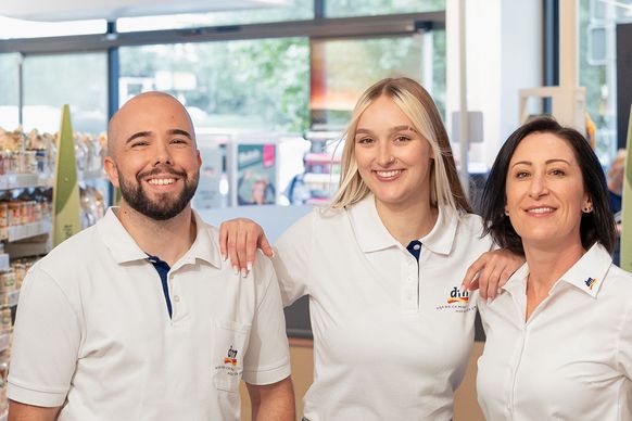 Drei lächelnde Angestellte in weißen Uniformen stehen in einem gut beleuchteten Ladengang, umgeben von Regalen mit verschiedenen Produkten.