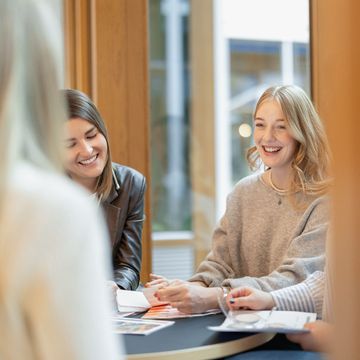 Zwei Frauen lächeln und unterhalten sich an einem Tisch in einem hellen Raum, mit Papieren und einer verschwommenen Person im Vordergrund.