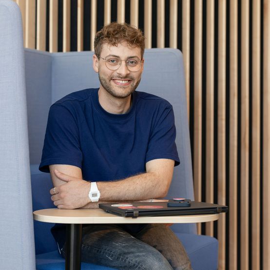 Mann in blauem T-Shirt sitzt vor einem holzverkleideten Hintergrund und lächelt in die Kamera