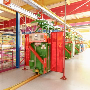 Eine farbenfrohe Industrieanlage mit Lichtkegel-Hängebahnen auf Schienen, grünen Geräten und roten Schutzwänden unter einer hellen Decke.