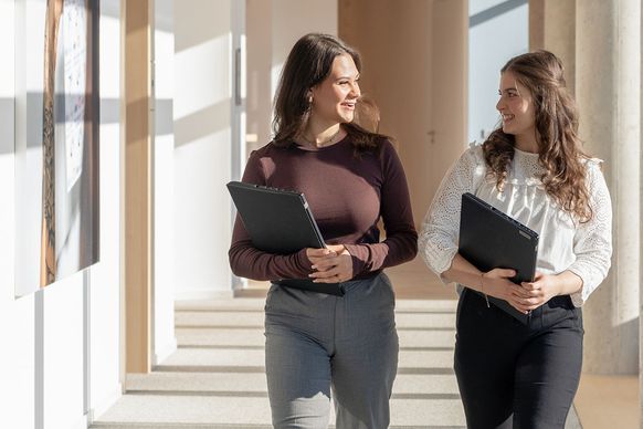 Zwei Frauen in legerer Geschäftskleidung gehen durch einen sonnenbeschienenen Flur, halten Aktenordner in der Hand und unterhalten sich.