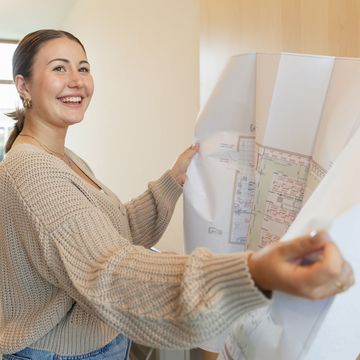 Lächelnde Frau, die in einem hellen Raum architektonische Entwürfe in der Hand hält und begutachtet. Sie trägt einen beigen Pullover und Reifenohrringe.