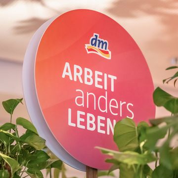 Ein rundes Schild mit Schriftzug „Arbeit anders Leben“ und einem dm-Logo, umgeben von Grünpflanzen.