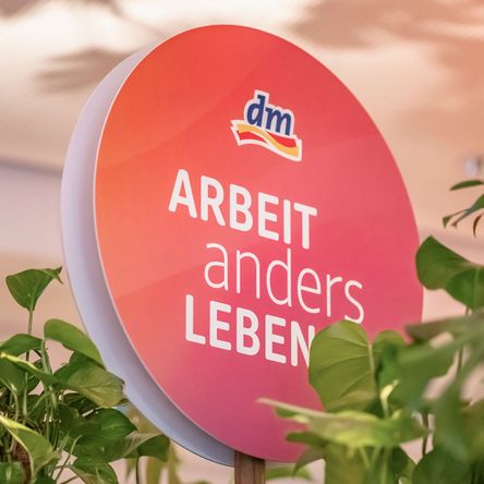 Ein rundes Schild mit Schriftzug „Arbeit anders Leben“ und einem dm-Logo, umgeben von Grünpflanzen.