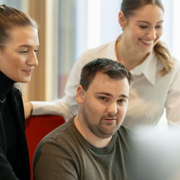 Drei Personen in einem Büro, zwei Frauen stehen und lächeln, ein Mann sitzt und schaut in die Kamera.