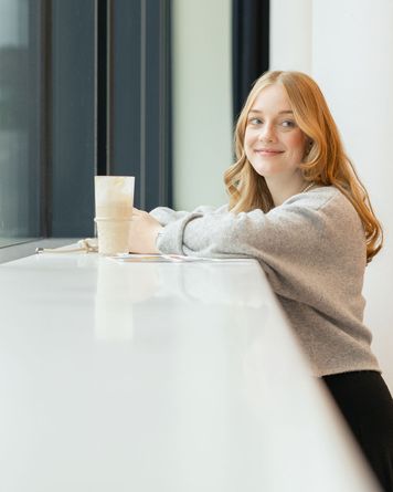 Frau mit langen blonden Haaren, die lächelnd an einem Fenster steht und einen großen Milchkaffee trinkt. Sie trägt einen grauen Pullover.