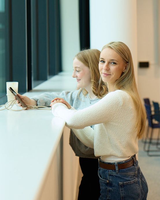 Zwei junge Frauen stehen an einem Fenster; die eine schaut auf ihr Telefon, die andere lächelt in die Kamera. Auf dem Tresen steht ein Getränk.