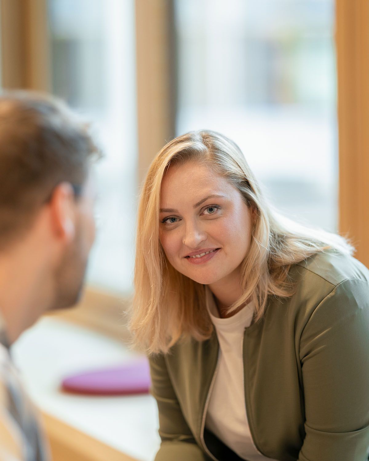 Frau mit blondem Haar, lächelnd, in grüner Jacke, blickt auf einen Mann mit verschwommenen Gesichtszügen im Vordergrund, in einem Innenraum.