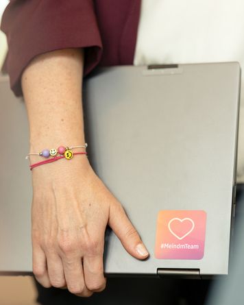 Eine Person hält einen Laptop mit einem „#MeindmTeam“-Aufkleber. Sie trägt ein Armband mit bunten Perlen, darunter auch Smiley-Gesichter.