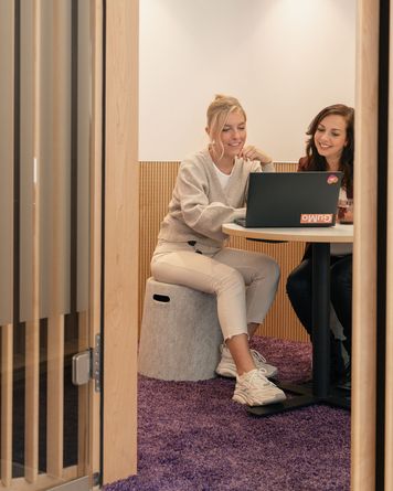 Zwei Frauen sitzen in einem kleinen, gemütlichen Raum mit Holzwänden und arbeiten gemeinsam an einem Laptop an einem runden Tisch mit einem lila Teppich.