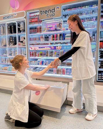 Angehende Drogistin reicht ihrer Kollegin Produkte aus einer Schublade vor einer Kosmetiktheke