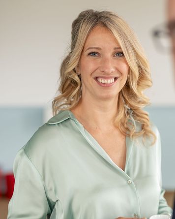 Lächelnde Frau mit blondem Haar in einer hellgrünen Bluse, die eine Tasse in der Hand hält, steht in einem Raum mit unscharfem Hintergrund.