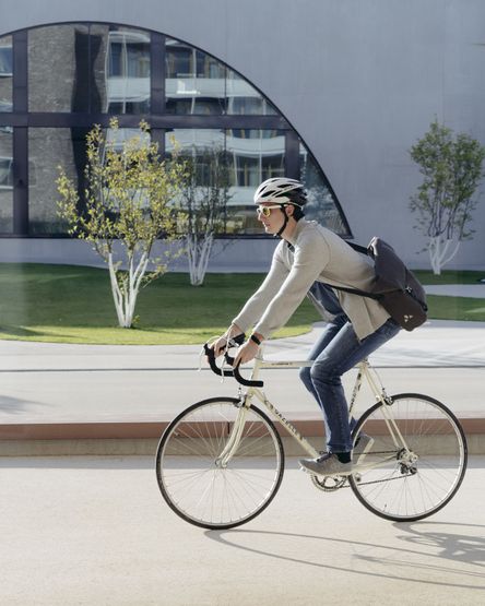 Eine Person, die einen Helm und eine Sonnenbrille trägt, fährt an einem sonnigen Tag Fahrrad, mit modernen Gebäuden und Bäumen im Hintergrund.