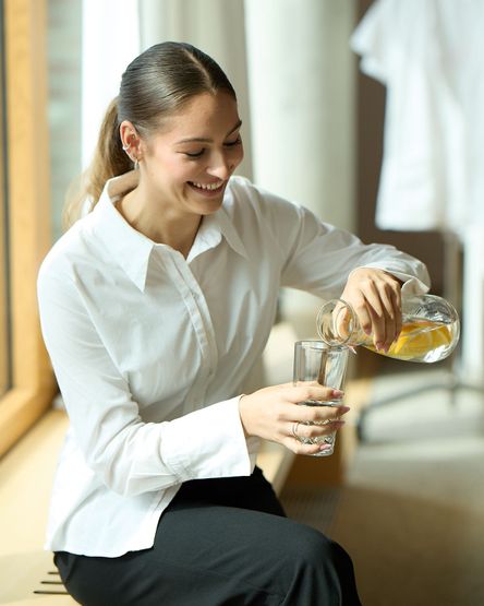 Eine lächelnde Frau in einem weißen Hemd gießt Zitronenwasser aus einer Glaskanne in ein Glas, während sie am Fenster sitzt.