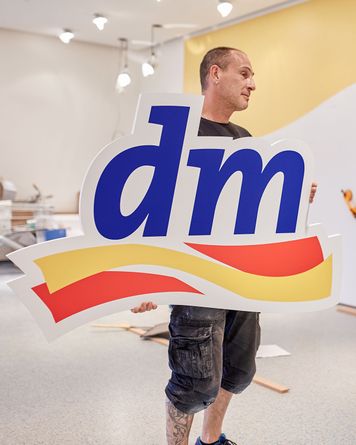 Mann mit großem „dm“-Logoschild mit blauen Buchstaben und roten und gelben Wellen in einem hellen Raum.