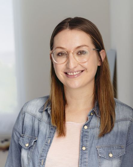 Frau mit Brille lächelt freundlich in die Kamera. Sie hat braune mittellange Haare und trägt ein offenes Jeanshemd.