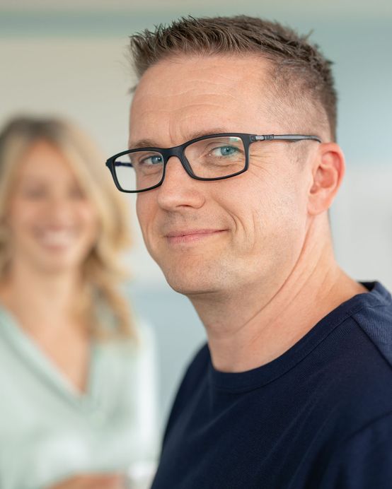 Mann mit Brille und kurzen Haaren, lächelnd, trägt ein marineblaues Hemd, mit einer unscharfen Person im Hintergrund.