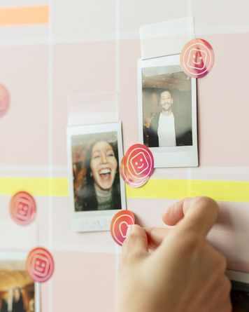 Hand platziert Smiley-Aufkleber neben Polaroid-Fotos auf einer Gittertafel. Ein Foto zeigt eine lachende Person, das andere eine Person mit einem breiten Lächeln.