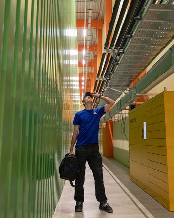 Eine Person in blauem Hemd und Mütze steht in einem hell erleuchteten Industriekorridor, schaut nach oben und hält eine schwarze Tasche in der Hand.