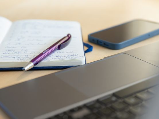 Notizblock mit lila Kuli neben Smartphone und Laptop