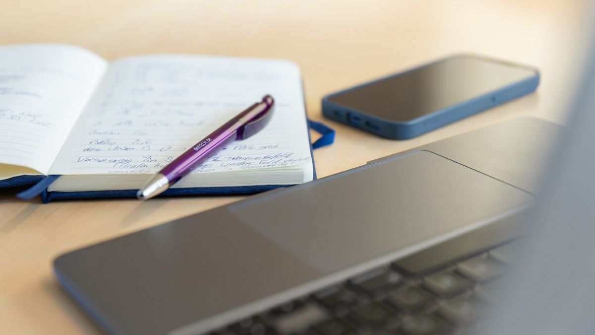 Notizblock mit lila Kuli neben Smartphone und Laptop