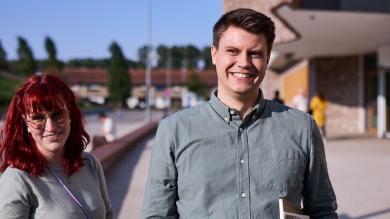 Ein lächelnder Mann und eine lächelnde Frau stehen an einem sonnigen Tag im Freien, mit einem unscharfen Hintergrund aus Bäumen und Gebäuden.