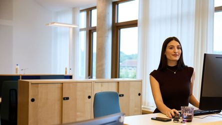 Eine Frau in einem schwarzen Kleid arbeitet an einem Computer in einem modernen Büro mit großen Fenstern und Holzschränken.