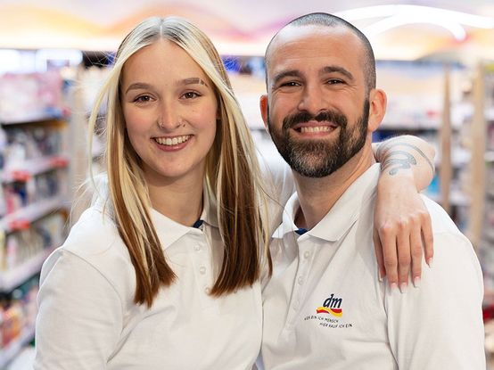 Zwei Verkäufer in weißen dm-Polo-Shirts lächeln in die Kamera. Im Hintergrund sind Warenregale zu sehen.