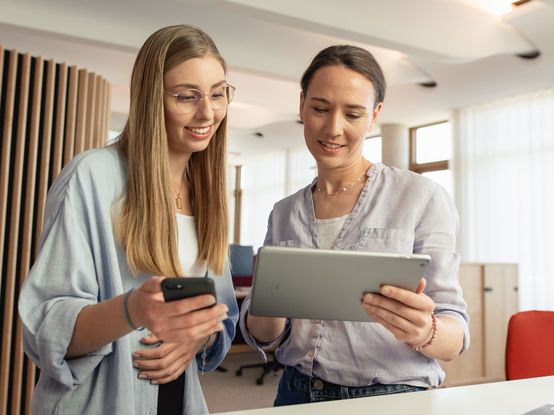 Angehende Kauffrau für Marketingkommunikation mit Ausbilderin gleichen Infos auf Smartphone und Tablet ab