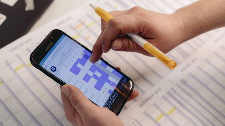 Hände, die ein Smartphone halten, das ein buntes Raster anzeigt, mit einem Stift, der auf den Bildschirm zeigt, vor einem Hintergrund mit Arbeitsplänen.