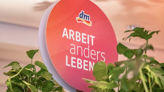 Ein rundes Schild mit Schriftzug „Arbeit anders Leben“ und einem dm-Logo, umgeben von Grünpflanzen.