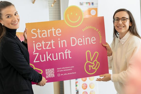 Eine lächelnde Person mit Brille hält ein farbenfrohes Schild mit Text und Grafiken in der Hand.