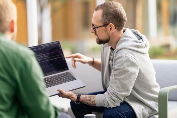 Ein Mann in einem grauen Kapuzenpullover zeigt einer anderen Person im Freien den Code auf einem Laptop und deutet dabei auf den Bildschirm.