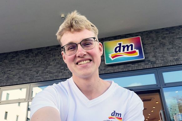 Eine lächelnde Person in einem weißen Hemd mit einem „dm“-Logo steht vor einem Gebäude mit einem „dm“-Schild über dem Eingang.