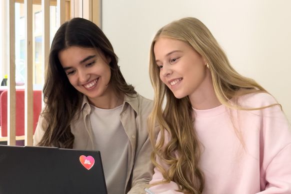 Zwei junge Frauen lächeln und schauen auf einen Laptop-Bildschirm in einem Innenraum. Die eine trägt ein beiges Hemd, die andere einen rosa Pullover. Auf dem Laptop befindet sich ein Herzaufkleber.