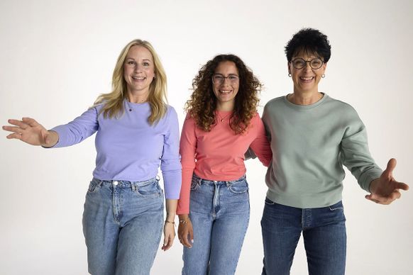Drei Frauen in legerer Kleidung stehen lächelnd beieinander und strecken einen Arm aus, vor einem schlichten weißen Hintergrund.