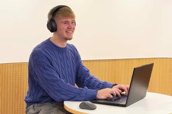 Mann in blauem Pullover und Kopfhörern, der lächelnd an einem kleinen runden Tisch auf einem Laptop tippt.