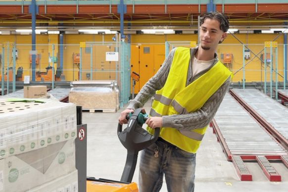 Ein Arbeiter in einem Lagerhaus, der eine gelbe Weste trägt, bedient einen Palettenhubwagen, um Waren zu transportieren, umgeben von Regalen und Lagerbehältern.