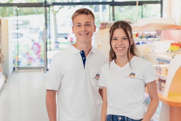 Zwei lächelnde Angestellte in weißen Hemden stehen in einem hell erleuchteten dm-Ladengang, mit Regalen voller Produkte im Hintergrund.