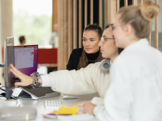 Drei Frauen arbeiten in einer modernen Büroumgebung an einem Schreibtisch mit einem Computer, wobei eine auf den Bildschirm zeigt.